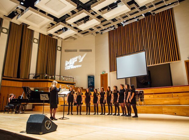 Choir of 10 girls singing with female conductor in large hall